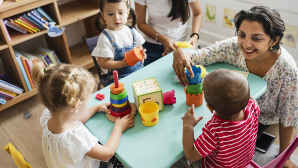 Children at nursery, sitting at table with teacher with stacking cups and other toys