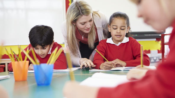 Primary school children sitting at tables in the classroom while the teacher helps one pupil