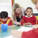 Primary school children sitting at tables in the classroom while the teacher helps one pupil