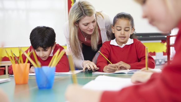 Primary school children sitting at tables in the classroom while the teacher helps one pupil