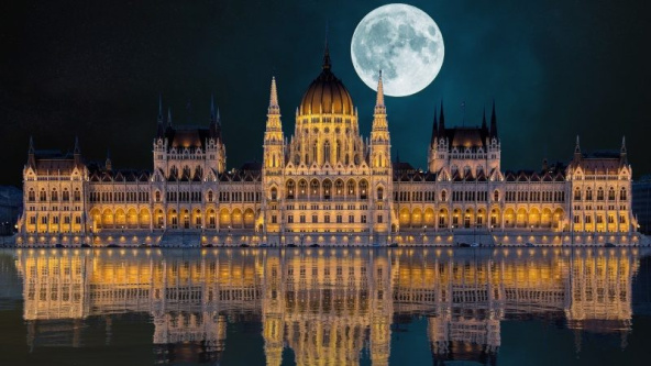 Scenic night view of The Hungarian Parliament Building in Budapest with a full moon illuminating the city skyline and historic architecture.