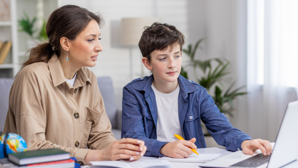 Tutor and teenage boy sitting at a desk in the living room, working together on school assignment