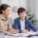Tutor and teenage boy sitting at a desk in the living room, working together on school assignment