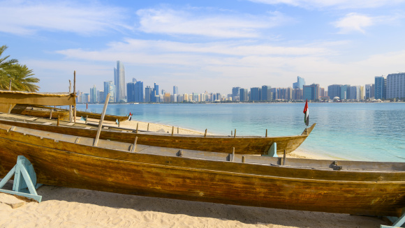 Wooden boats along a sandy beach on Marina Mall island with the modern skyline of Abu Dhabi across the water, in Abu Dhabi, United Arab Emirates