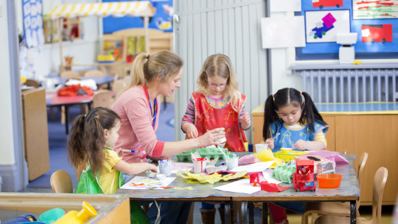 Children at nursery, sitting at table with teacher doing arts and crafts