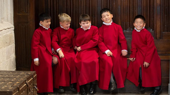 The boy choristers of Pilgrims' School, Winchester Cathedral, sit in a vestry wearing their red cassocks and white ruffs.