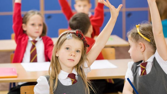 young girl in a prep classroom with a raised hand. Blurred children in the background with raised hands in red school uniform