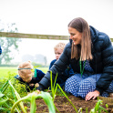 An adult woman with prep school children outside in a school garden planting vegetables in a planter box.