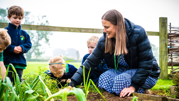 An adult woman with prep school children outside in a school garden planting vegetables in a planter box.
