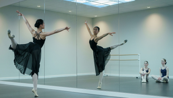 A young girl doing the pointe technique in a ballet class in front of a mirror. Two young girl sat in the background watching.