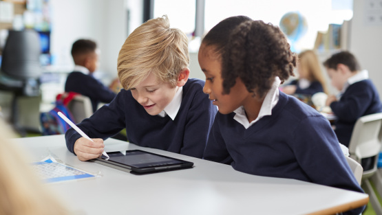 Two primary pupils - a girl and boy - sitting at a classroom table and working on a tablet