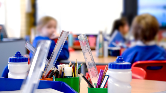 Primary school classroom with cups holding rulers, pencils and other stationery in the foreground and young pupils in the background