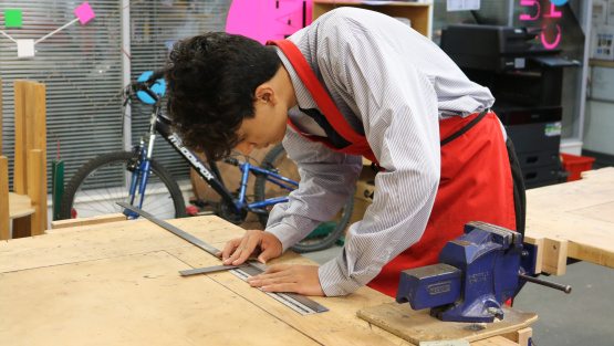 A boy in uniform and a red apron measures a length of metal on a work bench